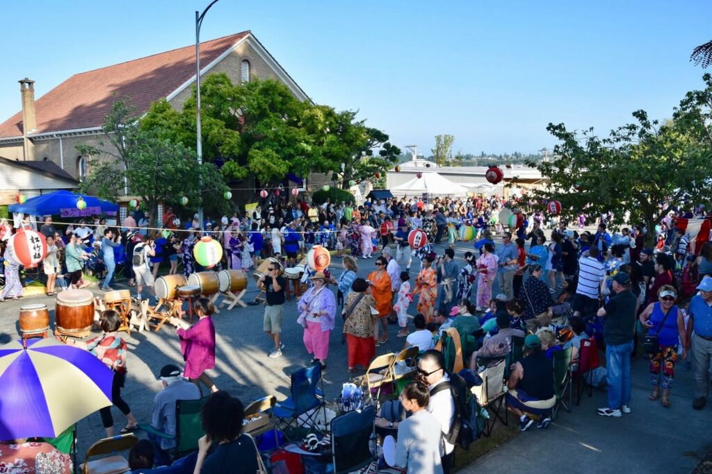 Busy outdoor festival with many people, colorful lanterns, and taiko drums in the foreground; attendees wear traditional yukatas and casual clothing under a clear blue sky.