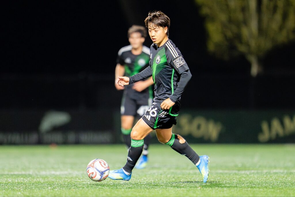 yu-tsukanome-1 - junglecity.com Soccer player in black and green kit dribbles a ball on a well-lit field at night with a teammate in the background