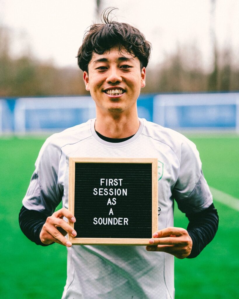 yu-tsukanome-3 - junglecity.com Smiling young man on a soccer field holding a sign that reads 'FIRST SESSION AS A SOUNDER'.