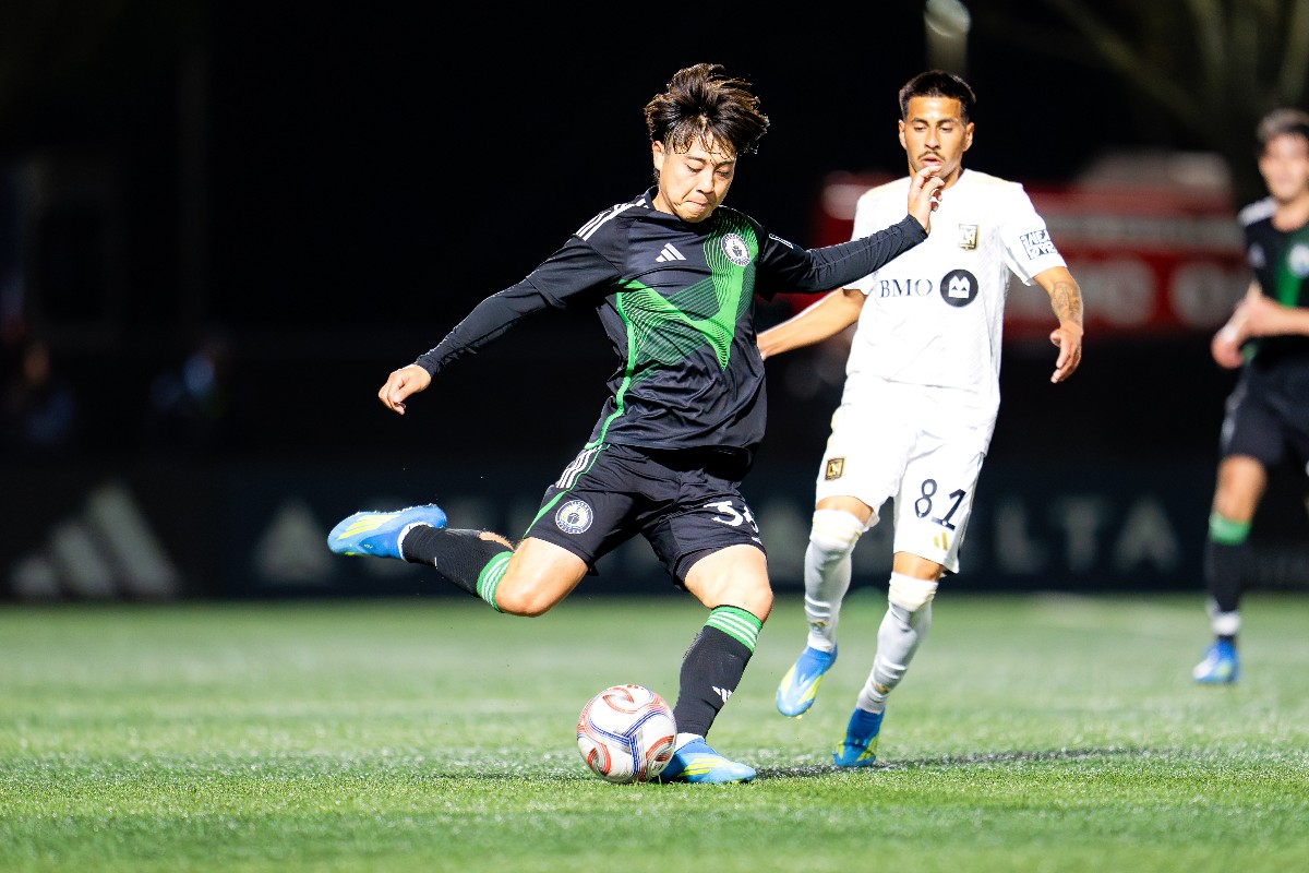 Soccer player in a black-green kit kicking a ball on a grass field during a night match, with a white-jersey opponent nearby.