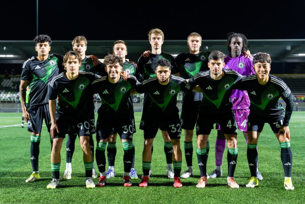 yu-tsukanome-6 - junglecity.com Soccer team lined up for a group photo on a night-green field, players in black and green jerseys with arms around shoulders.