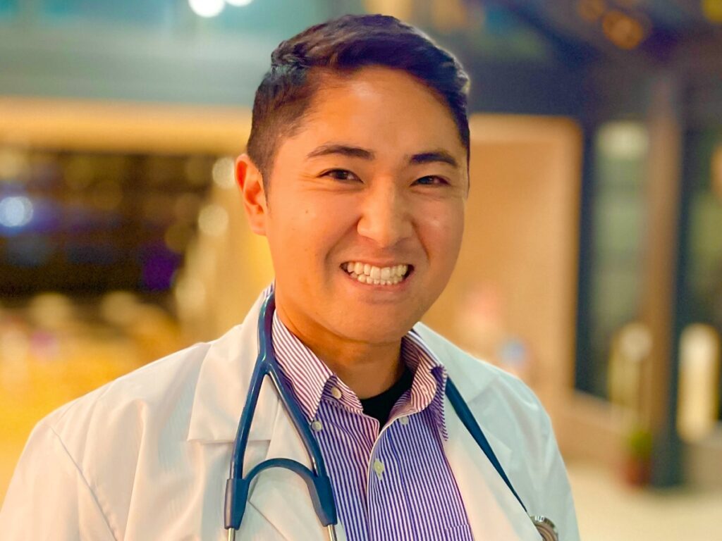 Smiling male doctor in a white lab coat with a stethoscope in a hospital corridor.
