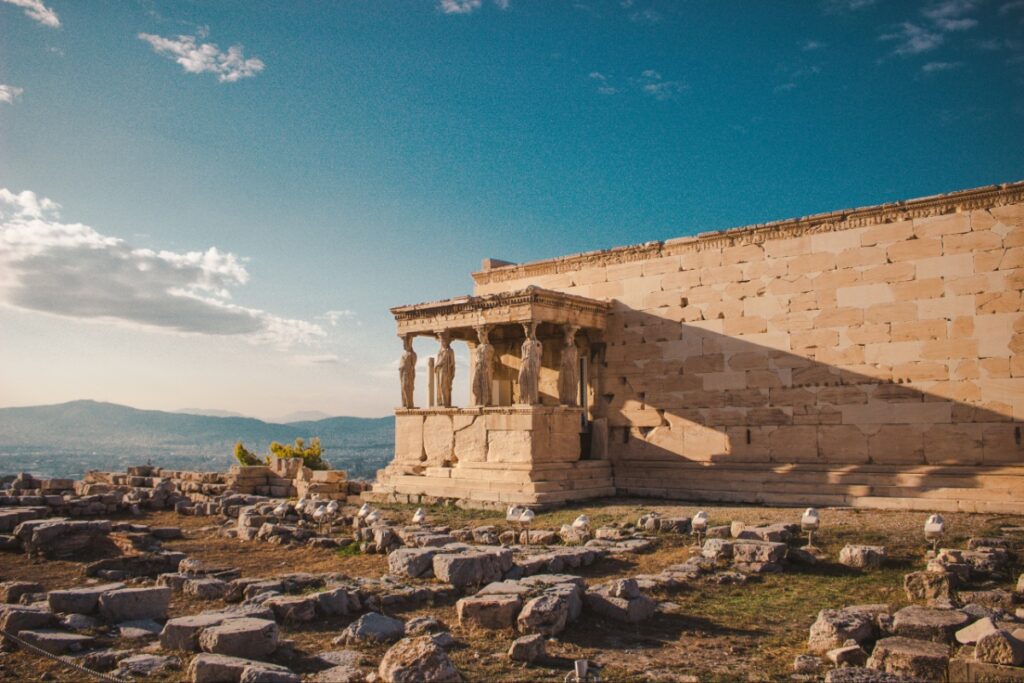 Ancient temple ruin with a row of statues on a columned porch, beside a long stone wall and scattered rocks under a blue sky.