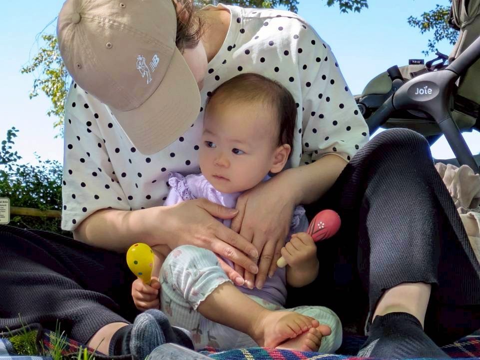 baby-massage-10 - junglecity.com Adult in a polka-dot shirt holding a baby on a blanket outdoors; a stroller is visible in the background.