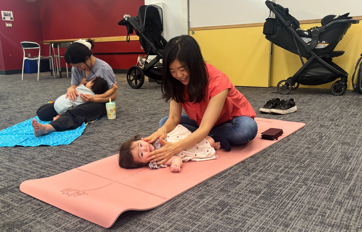 A caregiver smiles while gently massaging a baby lying on a pink mat in a class setting. A mother with a baby sits on a blue mat nearby.