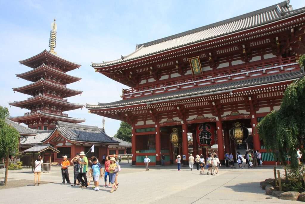 Temple complex with a multi-tiered pagoda and a large red gate, visitors walking on a sunny day.