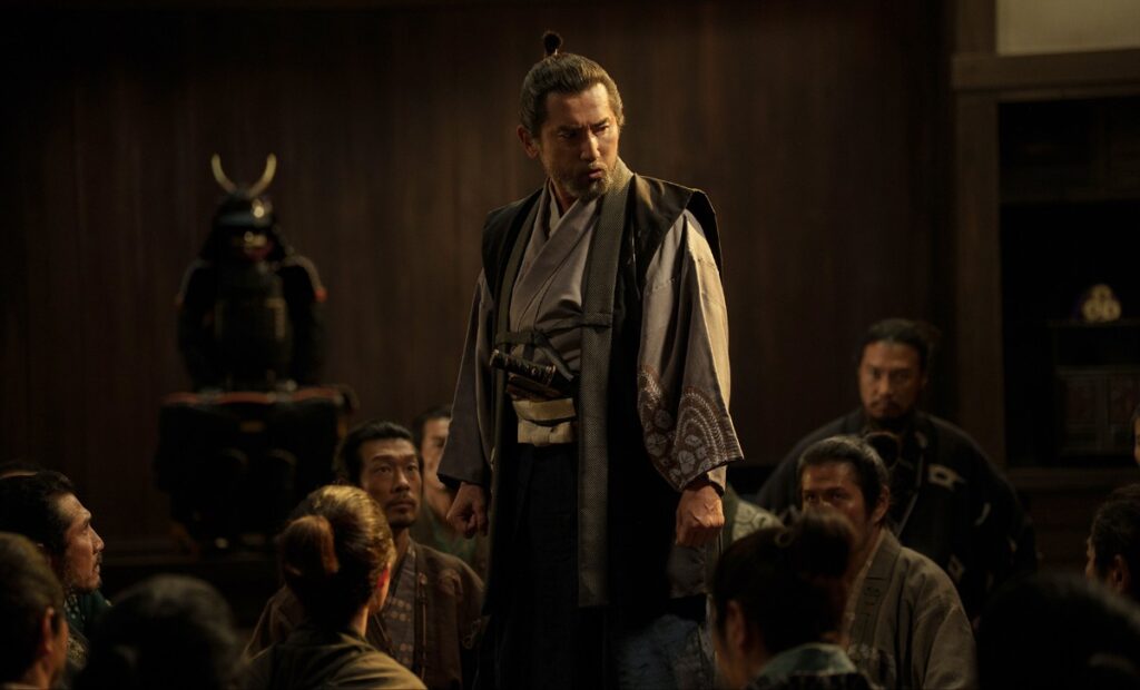 A man in traditional Japanese robes stands on a raised platform, addressing a seated group in a dim wooden hall with samurai armor in the background.