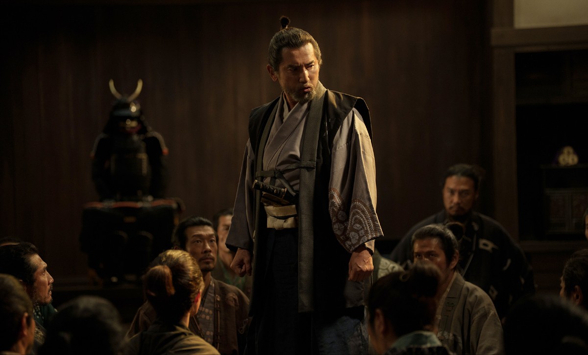 A man in traditional Japanese robes stands on a raised platform, addressing a seated group in a dim wooden hall with samurai armor in the background.