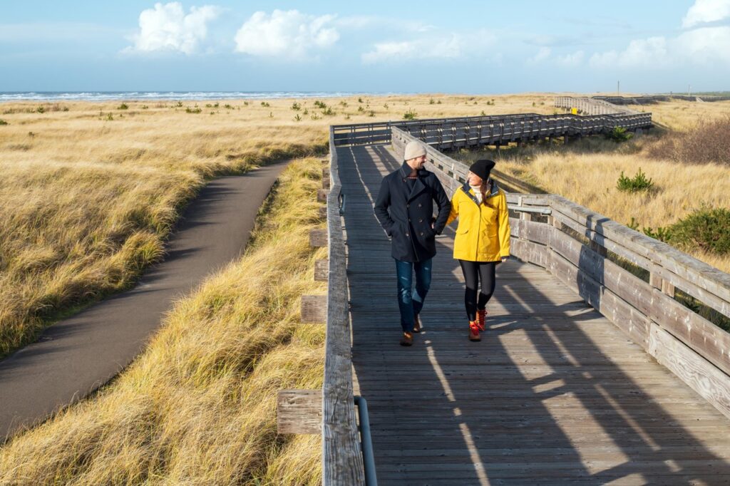 long-beach-3 - junglecity.com Two people walk along a wooden boardwalk through sandy dunes toward the sea on a sunny day, wearing coats and beanies.
