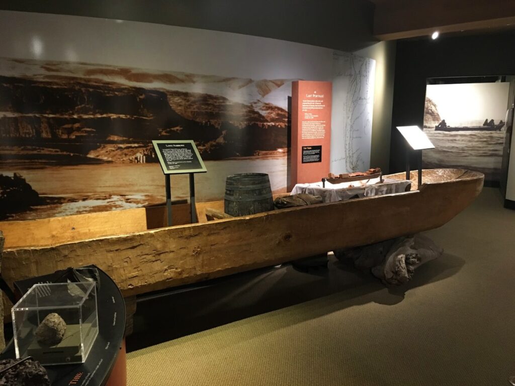 long-beach-5 - junglecity.com A large weathered wooden dugout boat displayed in a museum, with a barrel and display panels behind it and a rocky backdrop image on the wall.