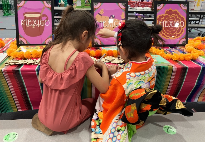 nanae-ohno-12 - junglecity.com Two girls in festive clothing sit at a Mexico-themed craft table, drawing with bright signs that say 'MEXICO' in the background.