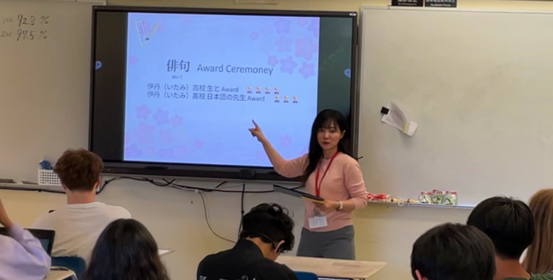 nanae-ohno-13 - junglecity.com Woman in a pink sweater stands at the front of a classroom, pointing to a slide on a large monitor that says 'Award Ceremony' during a presentation.
