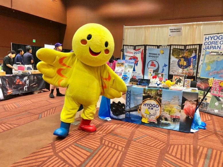 nanae-ohno-15 - junglecity.com Cheerful yellow mascot with outstretched arms posing at a convention booth featuring Hyogo Japan posters and banners.