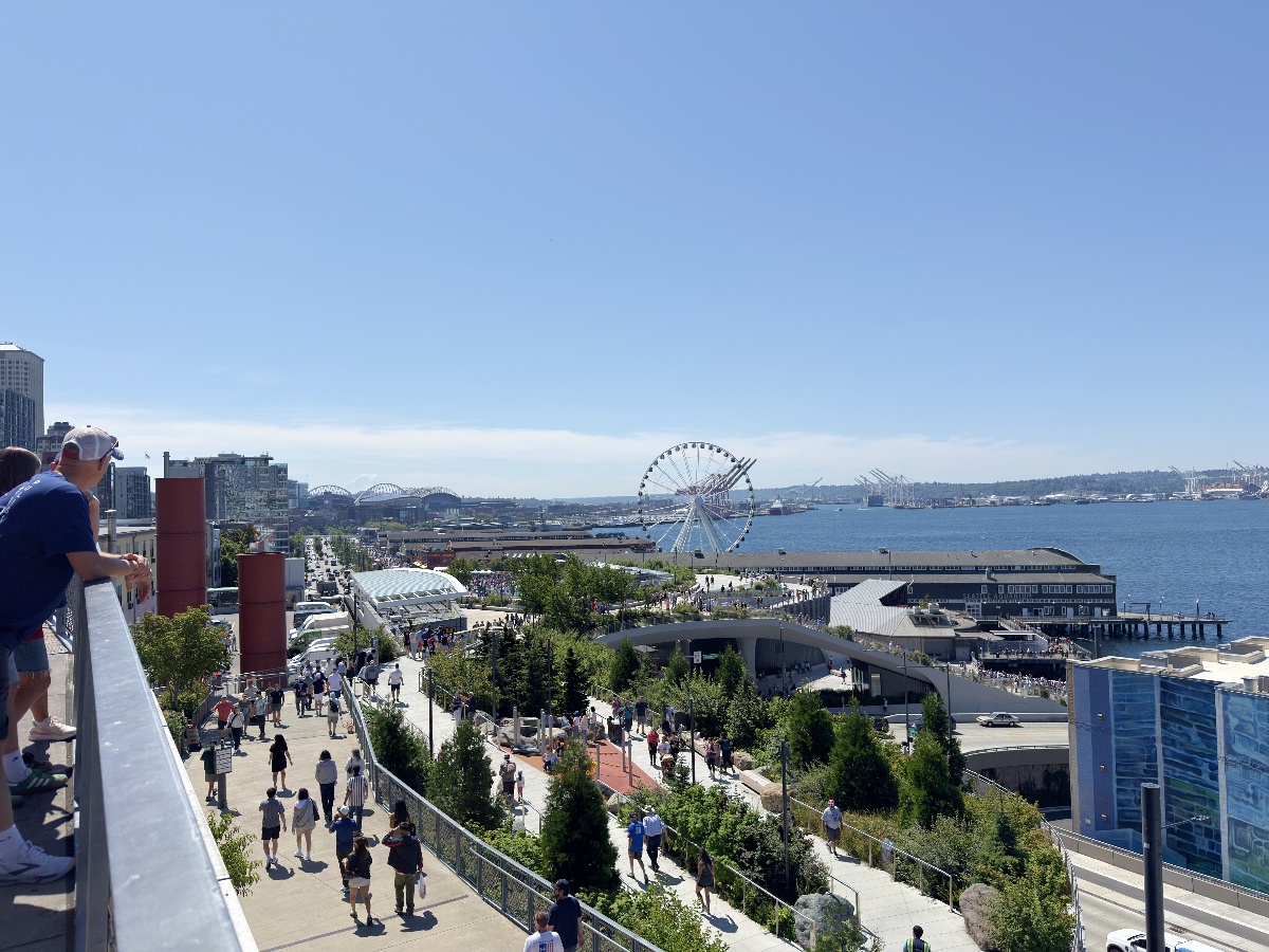 Crowded waterfront promenade with a Ferris wheel visible over the bay on a sunny day.