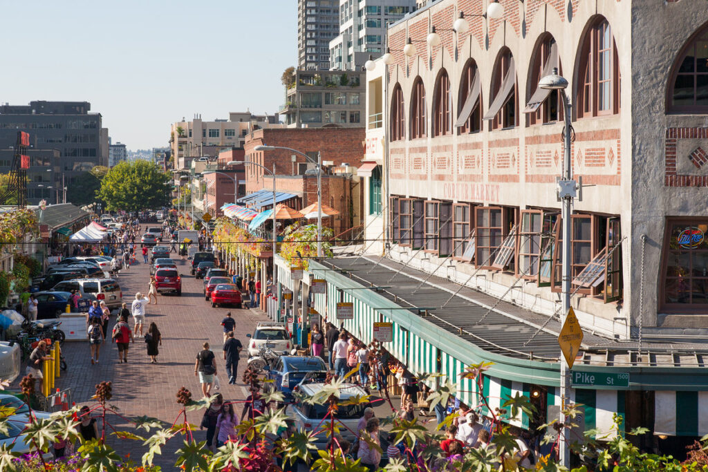 Busy city street market scene with striped awnings, pedestrians, and parked cars along brick storefronts on a sunny day.