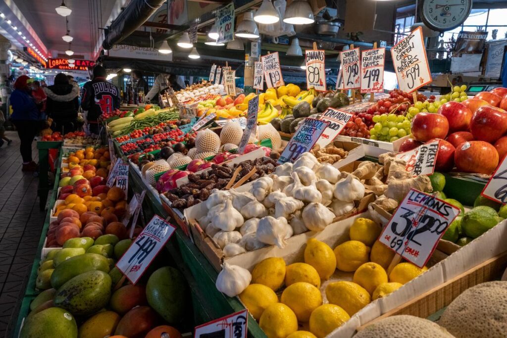 Bustling indoor fruit market with piled produce and handwritten price signs on stalls.