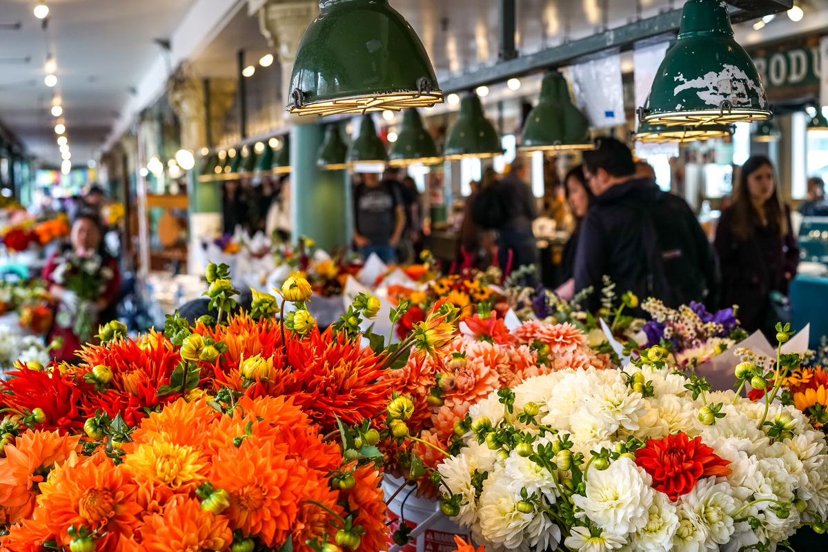 Indoor market flower stall with bright orange and white dahlias; shoppers browse in background under green industrial lamps.