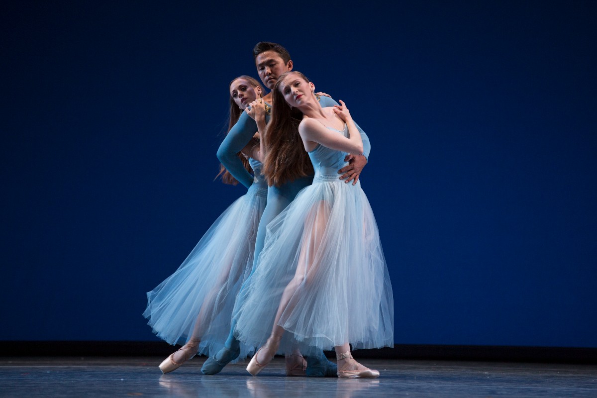 Three ballet dancers in light blue tutus performing a connected pose on a dark blue stage backdrop.