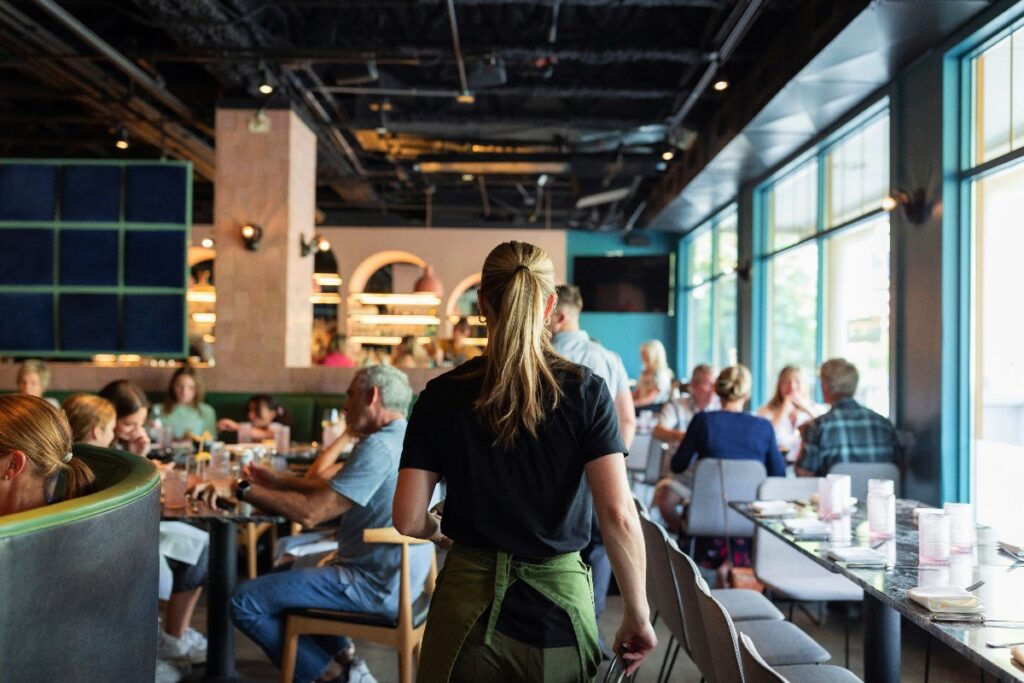 Server with ponytail, black shirt and green apron, walks through a busy restaurant seen from behind.