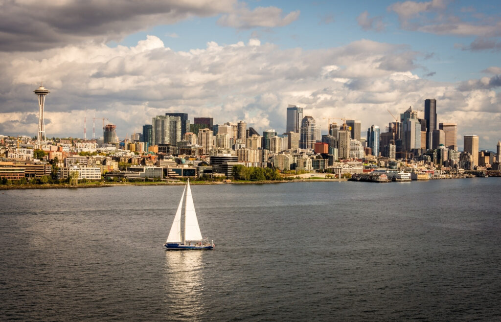 Sailboat on calm water with Seattle skyline and the Space Needle in the distance under a partly cloudy sky.