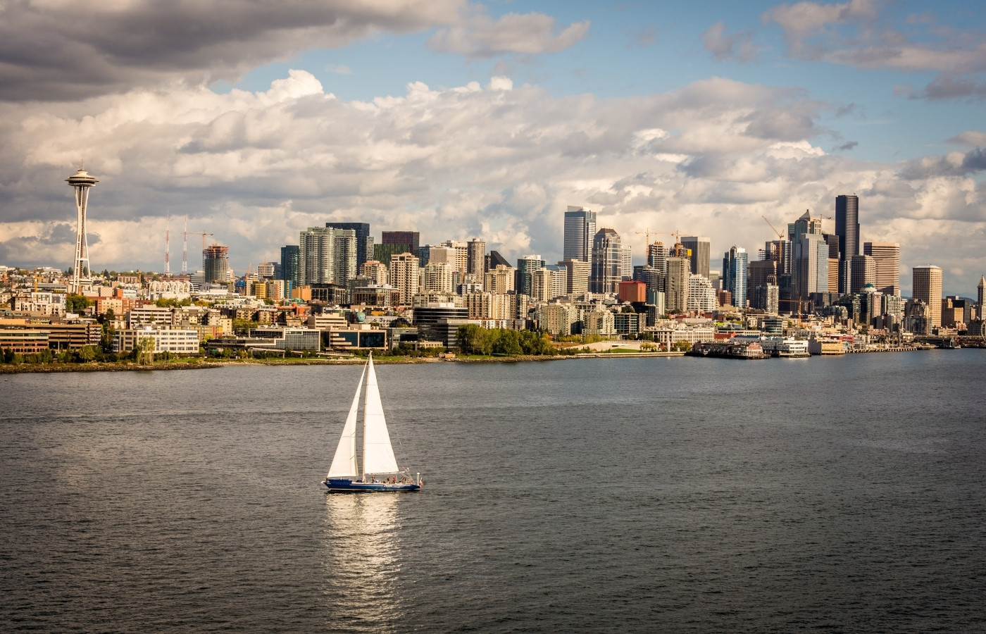 Sailboat on calm water with Seattle skyline and the Space Needle in the distance under a partly cloudy sky.