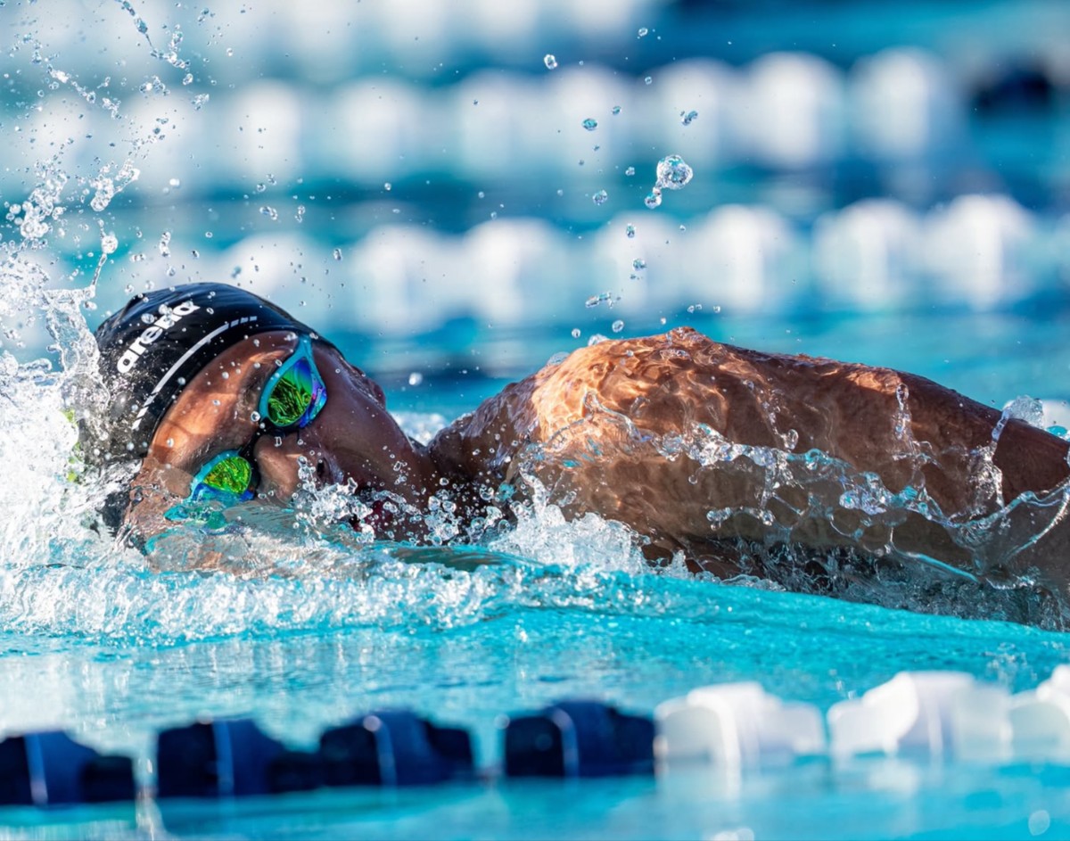 Swimmer wearing a black cap and green goggles glides through a blue pool with splashing water around her head in a freestyle stroke.