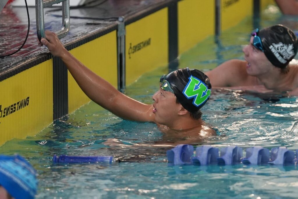 syunta-lee-2 - junglecity.com Swimmer in a black cap with a green 4 reaches for the pool ladder rail at the edge during a race by yellow lane markers and other swimmers nearby.