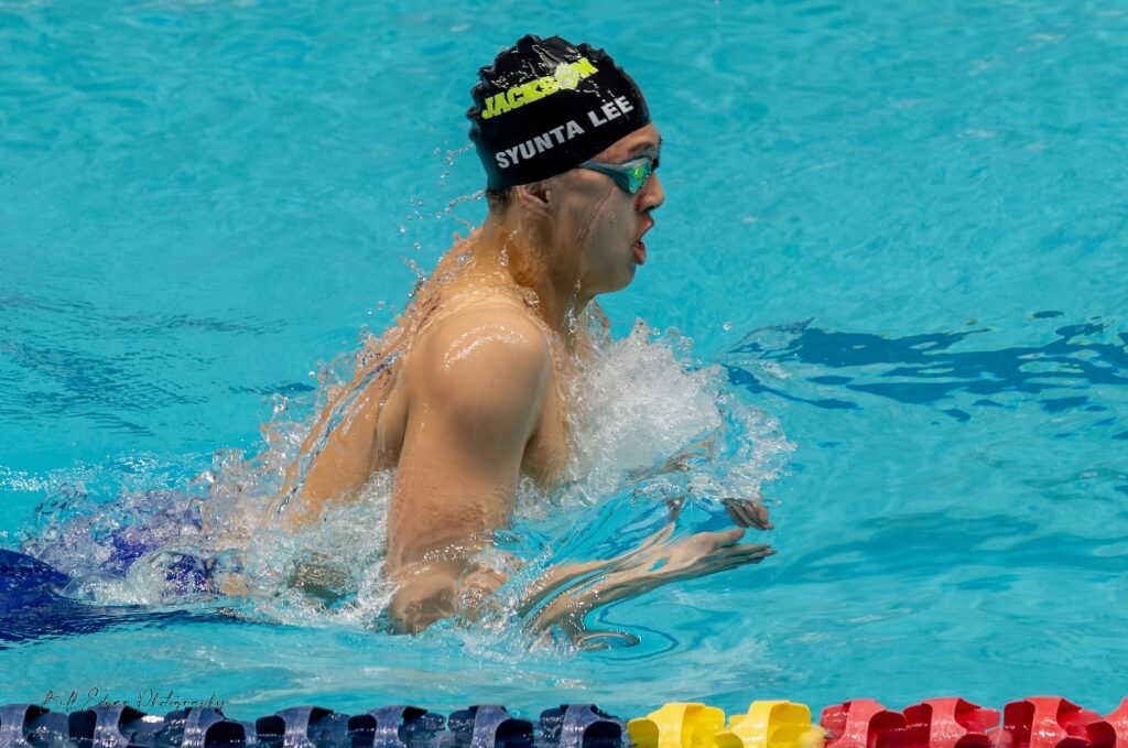 syunta-lee-3 - junglecity.com Swimmer in a black swim cap and green goggles gliding through a turquoise pool, creating a splash as their arms pull forward in a race stroke.