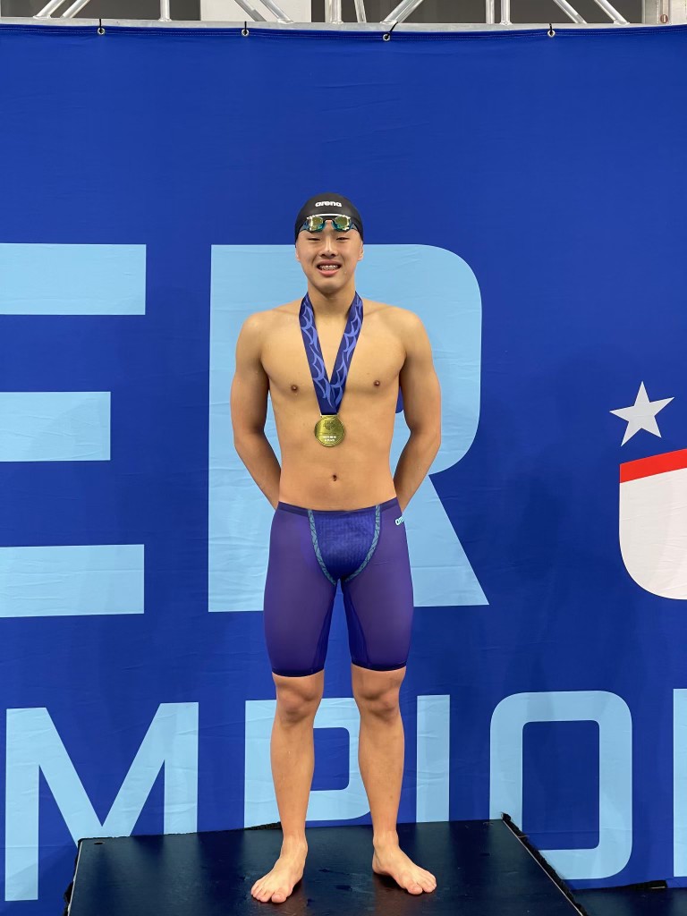 syunta-lee-4 - junglecity.com Male swimmer with gold medal on a purple swimsuit standing on a podium against a blue backdrop.