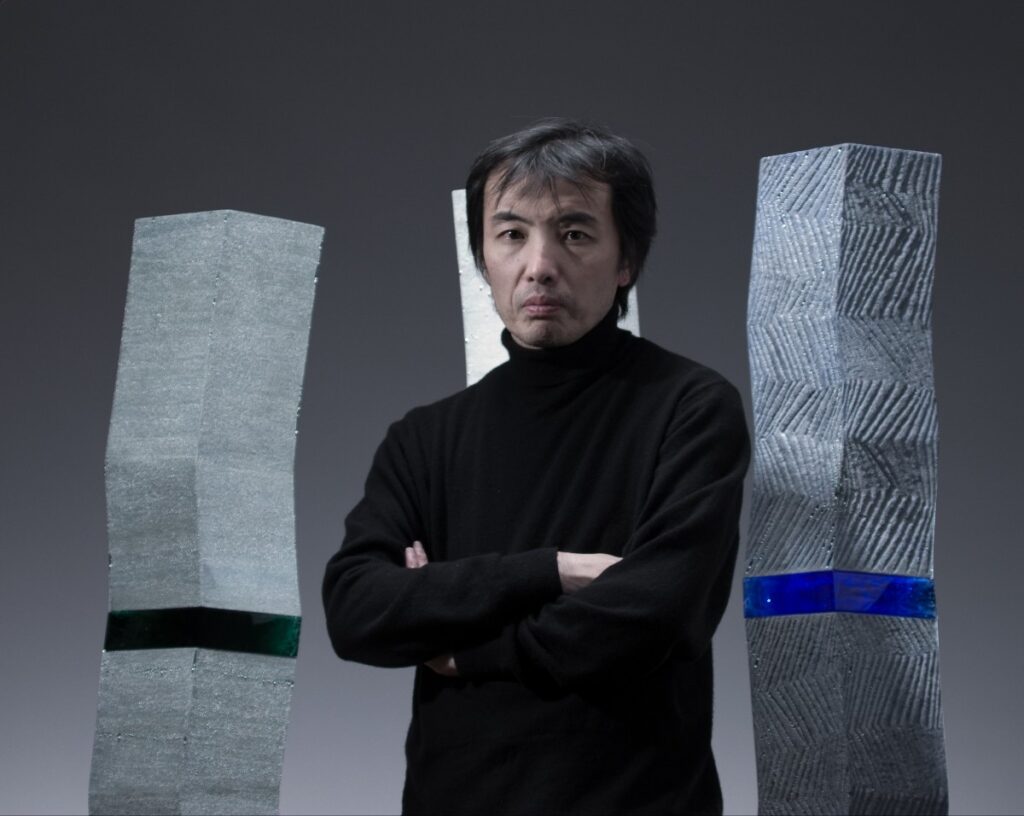 Man with arms crossed in a black turtleneck, posing between two tall metallic sculptures against a gray gradient backdrop.