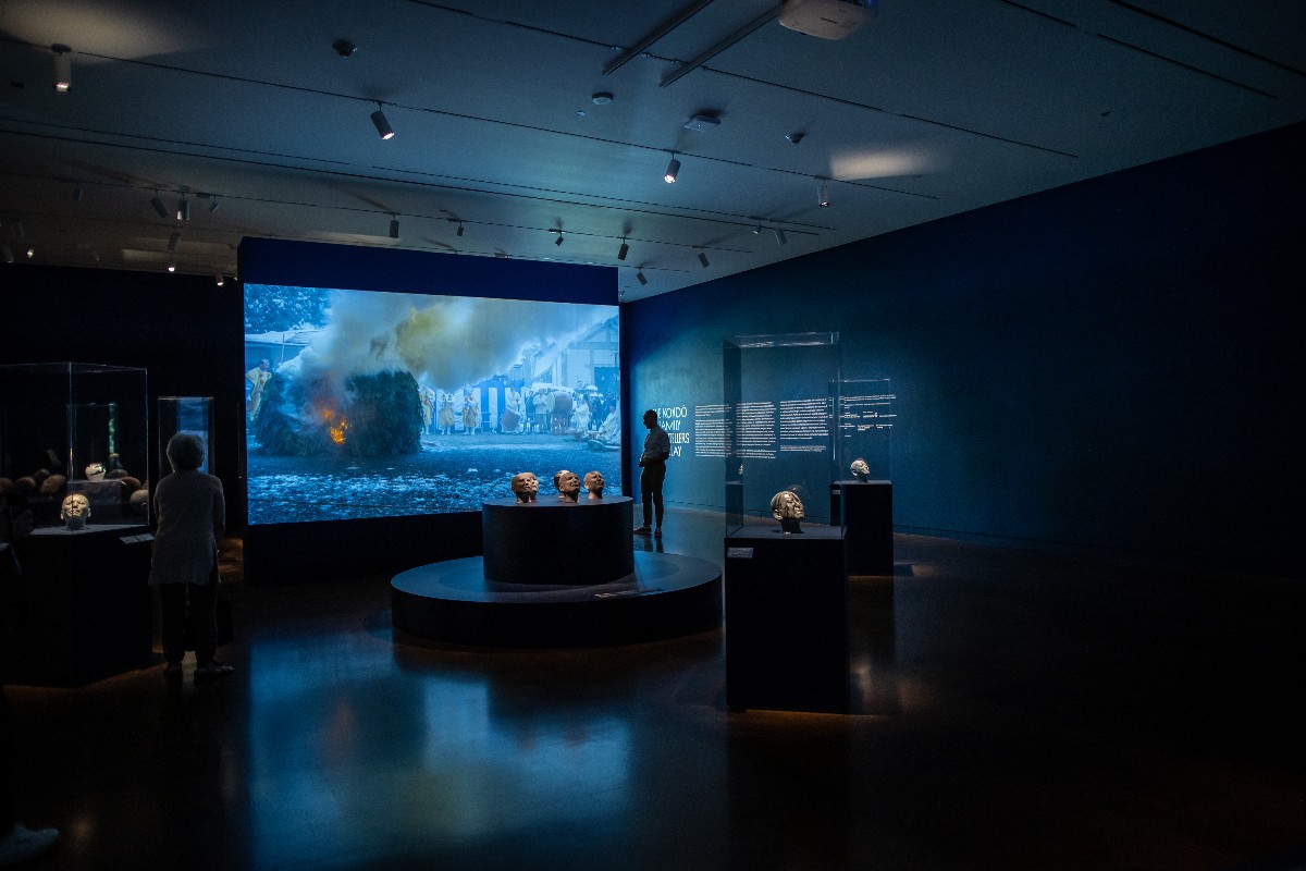 Dimly lit museum gallery with a large video wall showing a smoky street fire; busts on a circular pedestal in the foreground and visitors nearby.