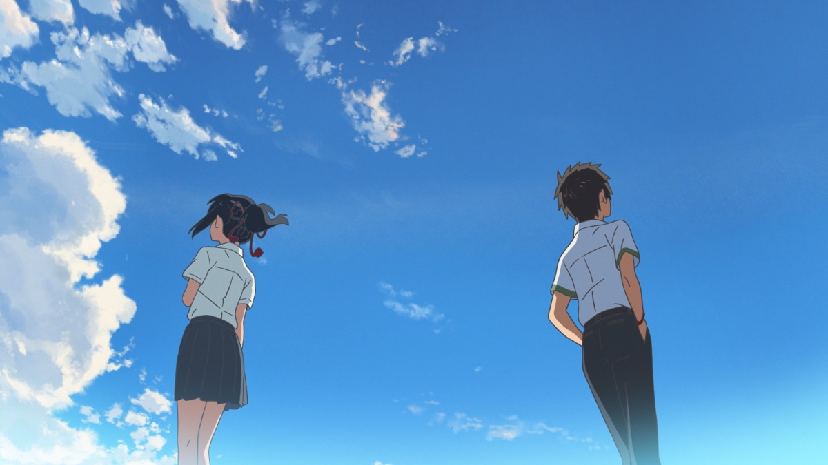 Two students in school uniforms stand apart, facing away, under a bright blue sky with scattered clouds.