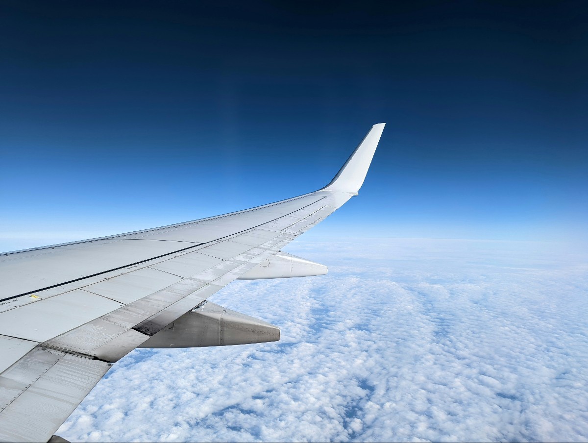 Airplane wing extending from the left toward the wingtip against a blue sky and cloud horizon, seen from a window.