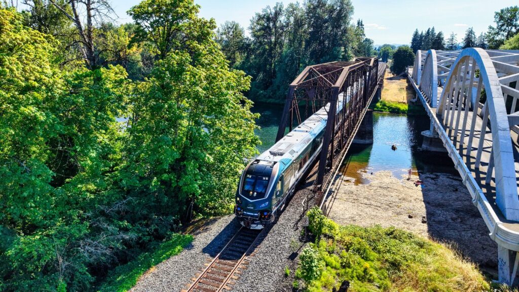 Passenger train crosses a dark steel truss bridge over a calm river, with a white arch bridge beside it and green trees along the bank.