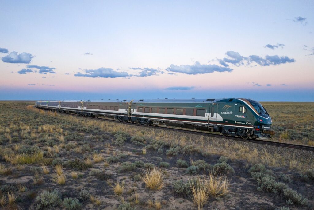 Green Cascades passenger train on a single-track in an arid prairie under a pastel sunset sky, stretching into the distance