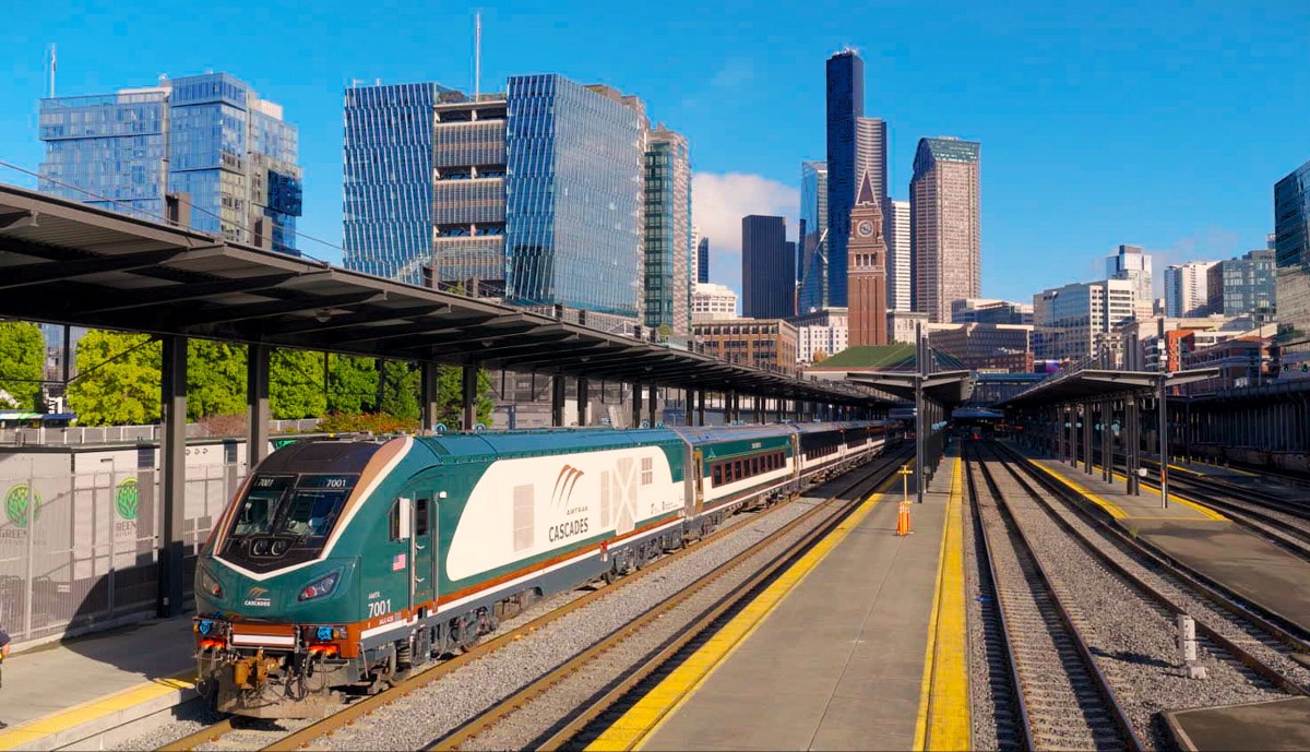 Amtrak Cascades train at a downtown station with glass skyscrapers in the background.