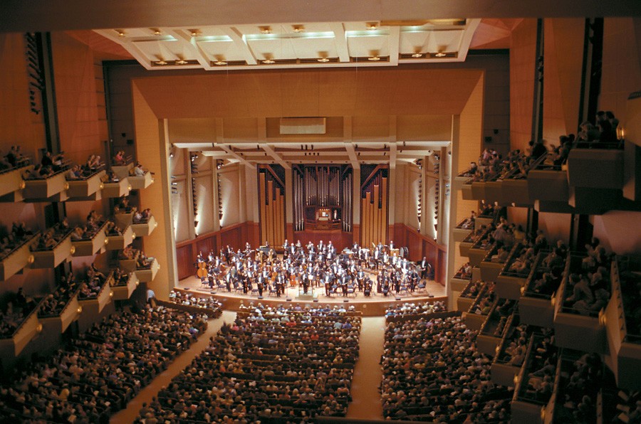 Concert hall viewed from the balcony with a full orchestra performing on stage and a large audience nearby.