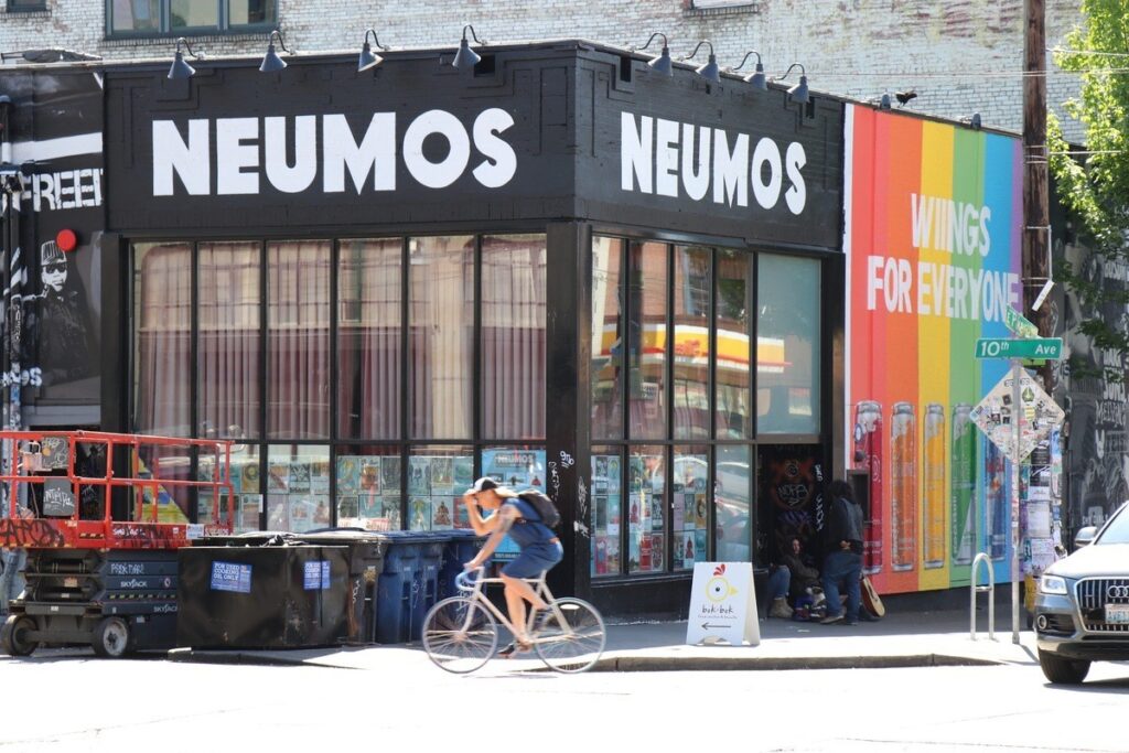 capitol-hill-21 - junglecity.com Exterior of Neumos venue with bold white signage and a rainbow mural that says'WINGS FOR EVERYONE' along the side wall. A cyclist rides by on the street.
