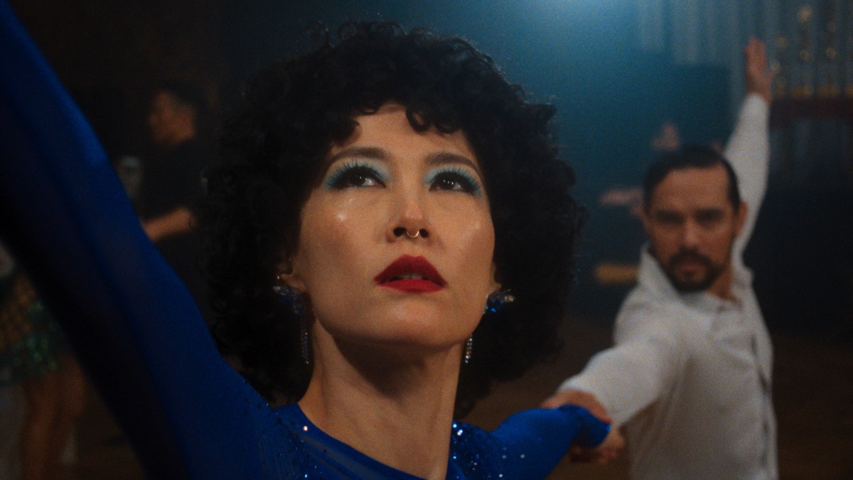 Close-up of a female dancer with blue eye makeup and red lipstick, looking upward while holding hands with a partner.