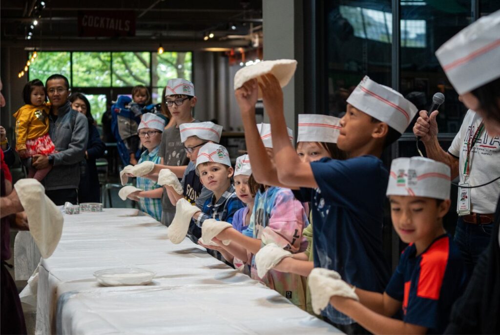 Group of kids wearing chef hats tossing dough at a long table during a pizza workshop, with adults watching in the background.