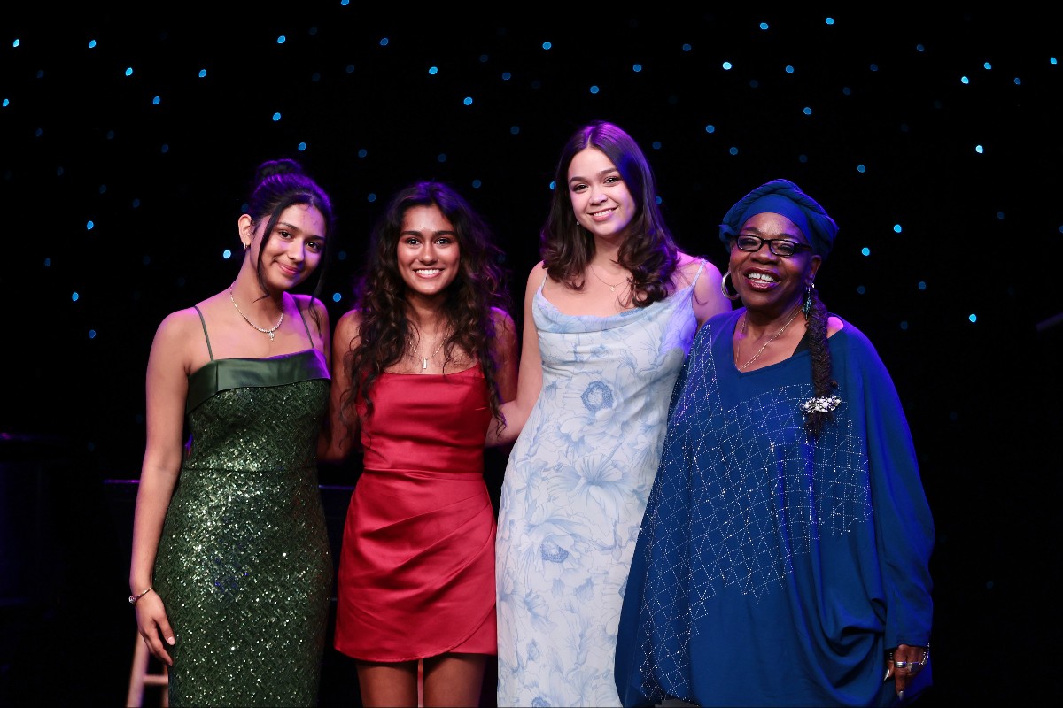 Four women stand arm-in-arm on a stage under blue fairy lights, smiling at the camera; left to right: a woman in a green glittery dress, a woman in a red satin dress, a woman in a light blue floral dress, and a woman in a blue outfit with a headwrap and glasses.