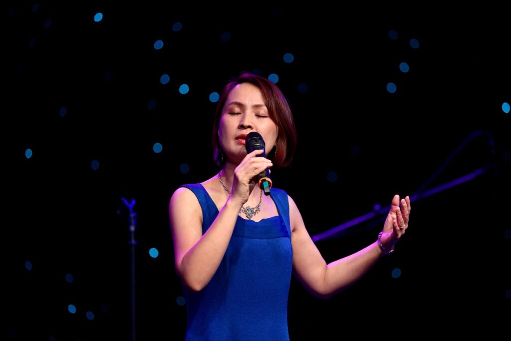 Woman in a blue dress sings into a microphone on stage, eyes closed, with blue bokeh lights in the background.