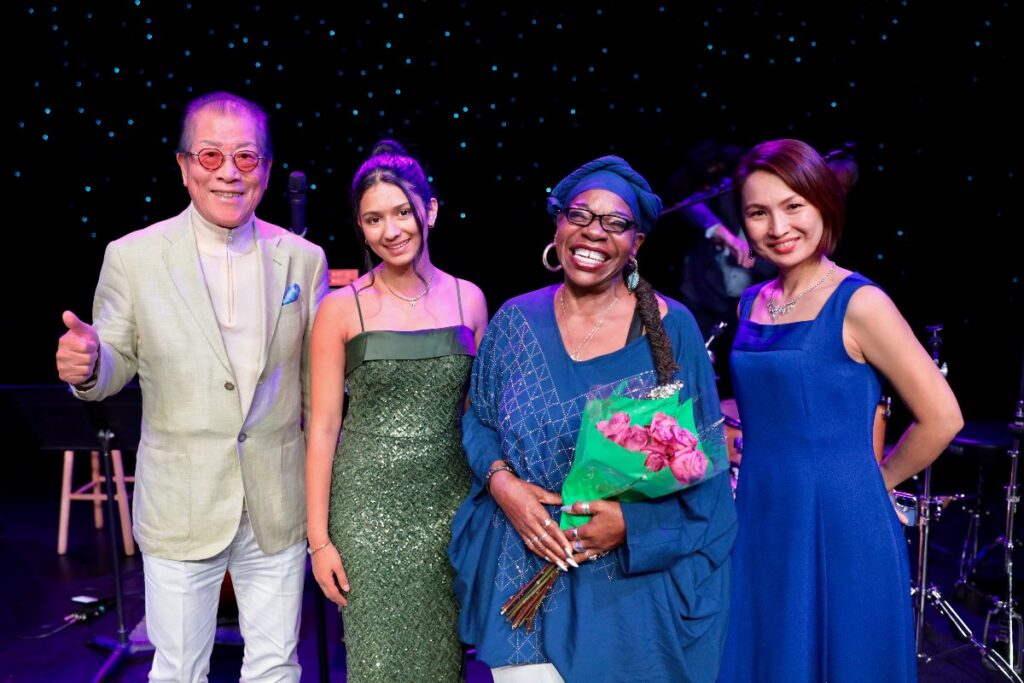 Four people pose on a dimly lit stage after a performance, smiling at the camera; a woman in blue holds a bouquet of pink roses in front.