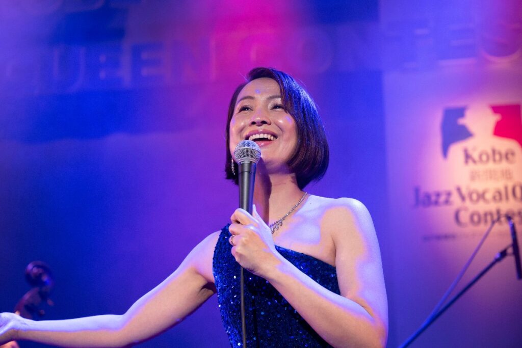 Female singer on stage in a blue sequined dress, smiling into a microphone under blue-purple stage lights; Kobe Jazz Vocal Contest banner in background.
