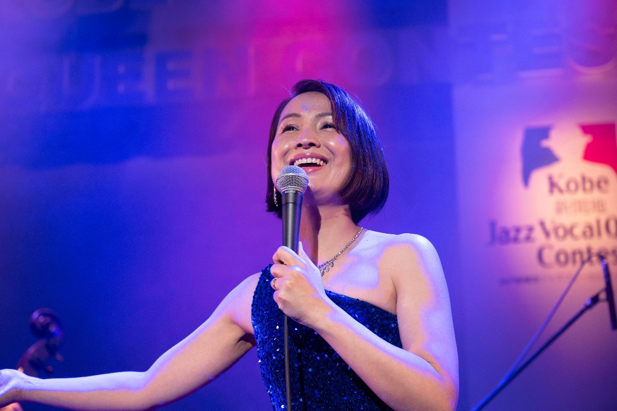 Female singer on stage in a blue sequined dress, smiling into a microphone under blue-purple stage lights; Kobe Jazz Vocal Contest banner in background.