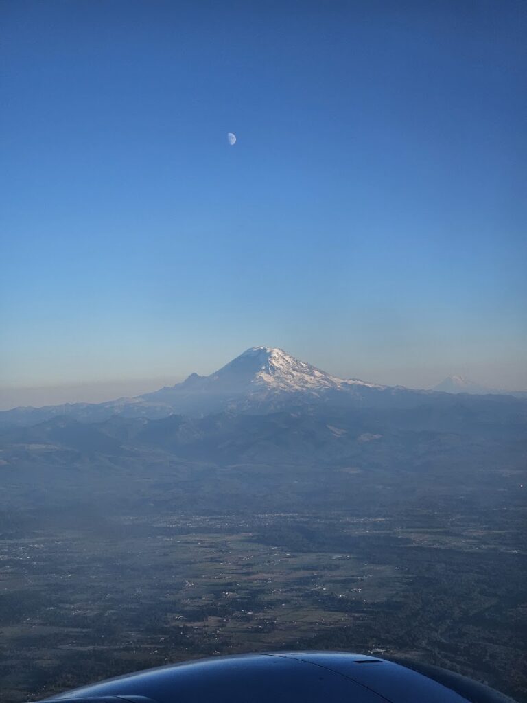 Snowy mountain peak seen from an airplane window with a crescent moon in a clear blue sky.