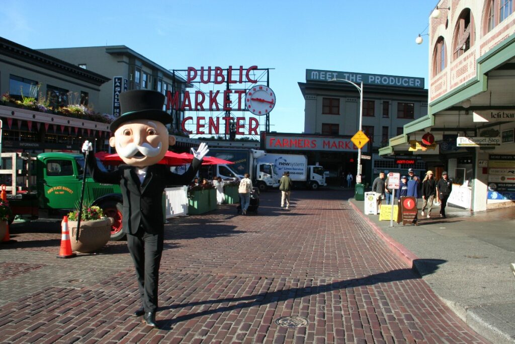 Mannequin in a tuxedo and top hat waving on a brick-paved street with 'Public Market Center' sign in the background and market stalls nearby