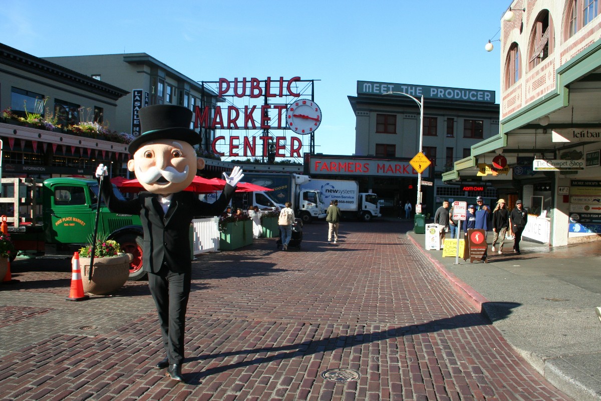 Mannequin in a tuxedo and top hat waving on a brick-paved street with 'Public Market Center' sign in the background and market stalls nearby