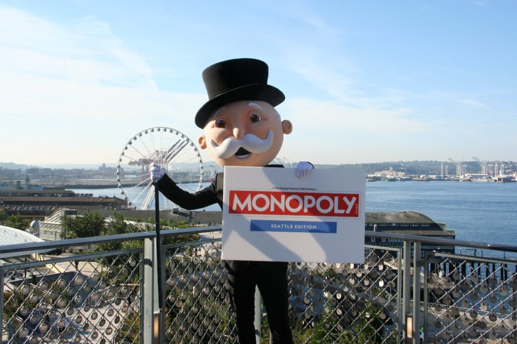 seattle-monopoly-2 - junglecity.com Mascot in a black top hat holding a large MONOPOLY Seattle Edition sign with a waterfront and ferris wheel behind.