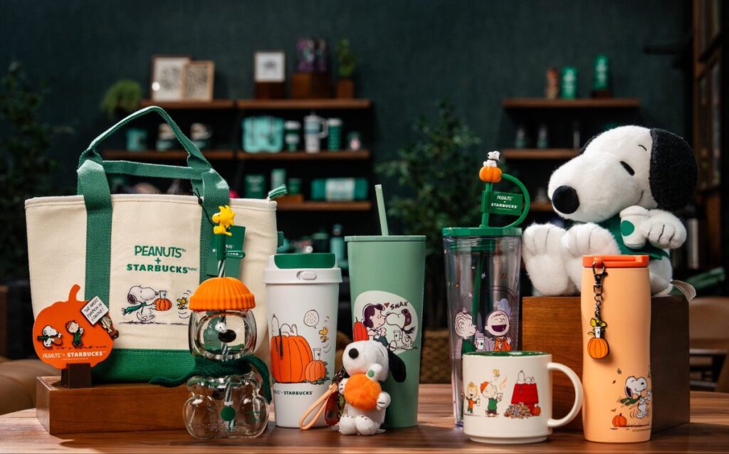 Display of Peanuts x Starbucks merchandise on a table: tote bag, tumblers, cups, and a Snoopy stuffed toy with pumpkin-themed accents.